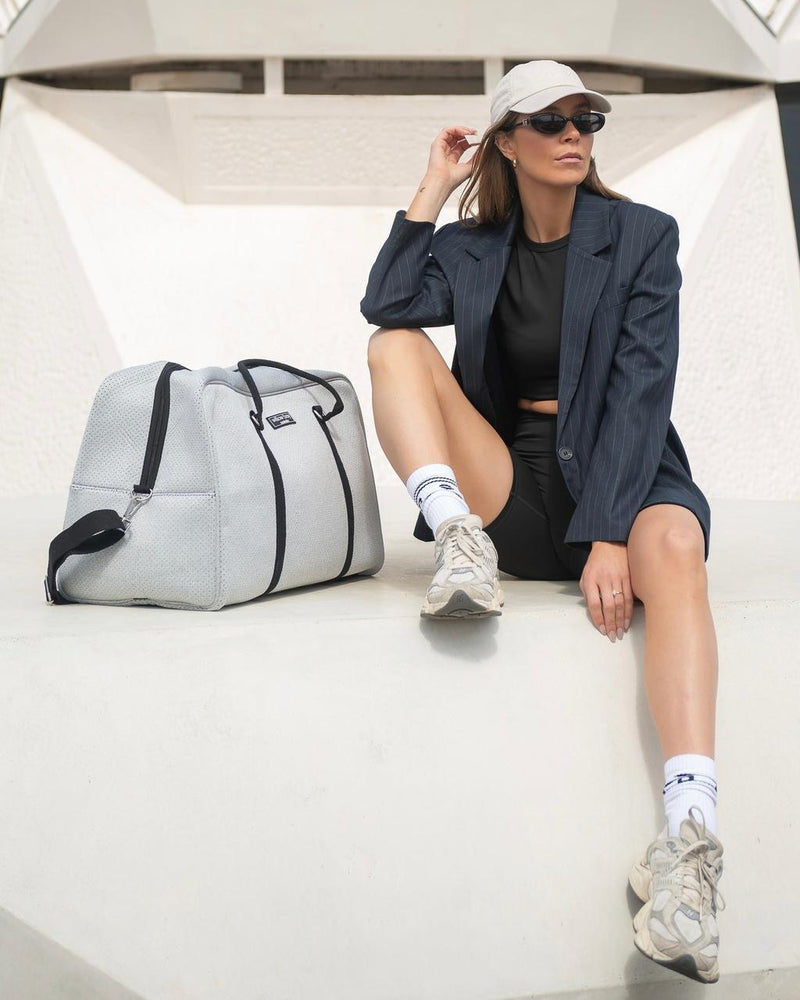 Stylish woman with a light marle neoprene Weekender Bag, featuring black straps, sitting on a white concrete structure.