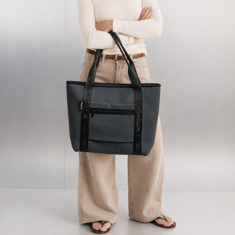 Woman holding a charcoal neoprene travel tote with black branded straps and a front zip pocket.