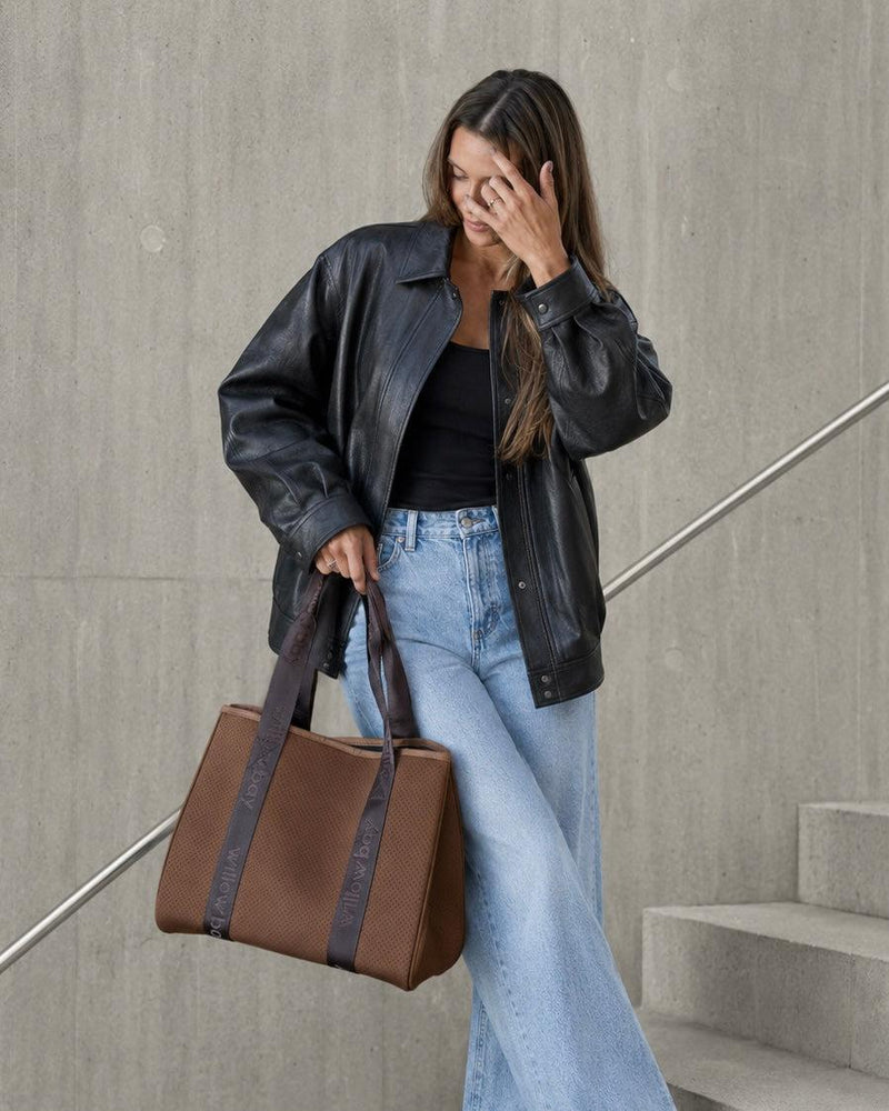 Woman in black leather jacket and jeans holding a rust brown neoprene tote with Willow Bay branded straps.